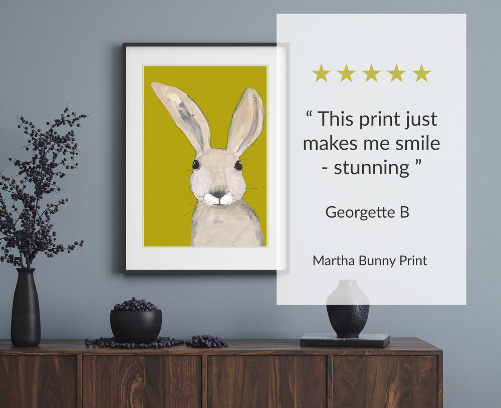 A framed art print of a bunny on a mustard yellow background, displayed on a grey wall above a wooden cabinet. A white overlay on the right features a five-star rating and a quote: 'This print just makes me smile - stunning' by Georgette B, Martha Bunny Print.